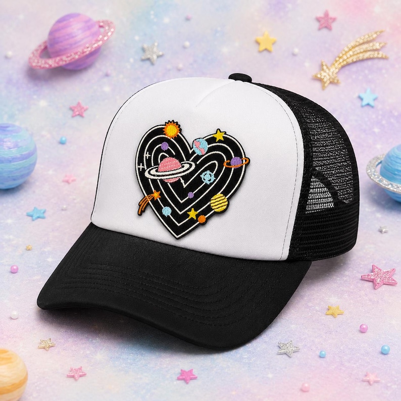 May include: A black and white trucker hat featuring a central embroidered patch of a black heart shape filled with white concentric lines and colorful celestial bodies including planets, stars, and a comet. The hat has a solid black brim and mesh back.