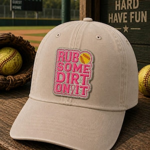 May include: A tan baseball cap with a pink and yellow patch that reads "RUB SOME DIRT ON IT". The cap is sitting on a wooden surface with a baseball glove and baseball in the background. The words "PLAY HARD HAVE FUN" are visible on a wooden sign.