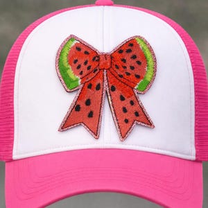 May include: A pink and white trucker hat featuring a watermelon bow applique. The bow has a red center with black seeds and green rind, outlined with glitter. The hat has a pink brim and mesh back.