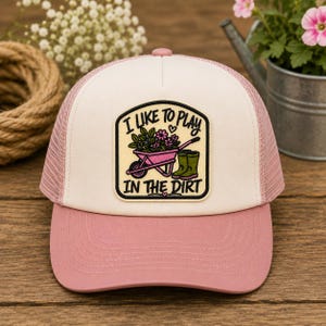 May include: A cream and dusty pink trucker hat featuring a central patch with a pink wheelbarrow, green boots, and flowers. The patch includes the black text I LIKE TO PLAY IN THE DIRT. The hat has a solid front panel and a breathable mesh back.