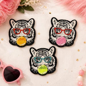 May include: Three embroidered tiger patches featuring white and black fur, red or teal glasses, and round bubblegum spheres in yellow, pink, or lime green. Each patch has a black glittery border and depicts a tiger head facing forward.