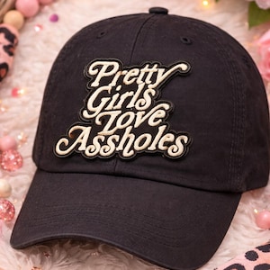 May include: Black baseball cap with the embroidered phrase "Pretty Girls Love Assholes" in a cursive font. The hat is made of a soft fabric and has a curved brim. The background includes pink flowers and decorative beads.