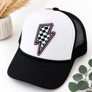 May include: A trucker hat featuring a white front panel with a black and white checkered lightning bolt patch outlined in pink and teal. The hat has a solid black brim, a black mesh back, and a black cord detail across the base of the front panel.