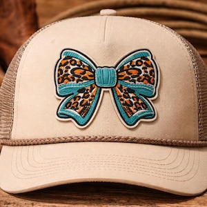 May include: Tan trucker hat with a brown mesh back and a decorative bow. The bow features a turquoise outline, a tan center, and a leopard print design in orange and brown. The hat has a curved brim.