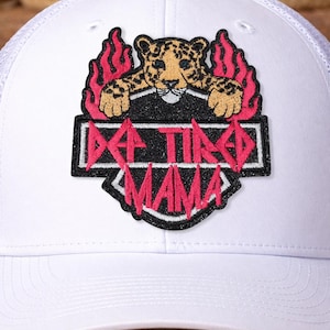May include: White trucker hat with a mesh back and a curved brim. The front features a patch with a leopard design, flames, and the text "DA TIRE MAMA" in pink.