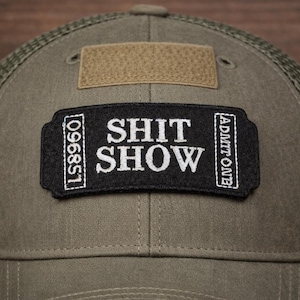 May include: Olive green trucker hat with a mesh back and a curved brim. The front features a black patch with the words "SHIT SHOW" in white, along with the text "ADMIT ONE" and the number "096851".