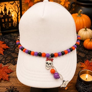 May include: A white baseball cap featuring a decorative beaded band in alternating orange, purple, and black colors. A silver-toned charm of a skull wearing a red bow hangs from the band, accompanied by a small bat charm and additional beads.