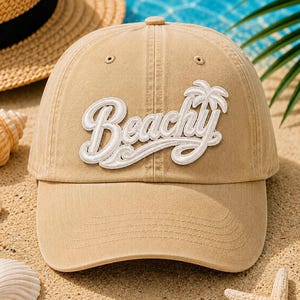 May include: Tan baseball cap with the word "Beachy" in white script and a palm tree graphic. The hat is on a sandy surface with a straw hat and seashells in the background. Perfect for a day at the beach.