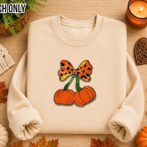 May include: A beige crewneck sweatshirt featuring a central patch of two orange pumpkins with green stems topped by a single large bow with an orange and black leopard print pattern. The design is centered on the front of the garment.