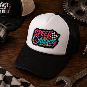 May include: A black and white trucker hat featuring a patch with the text SPEED ADDICT in pink and blue, a speedometer icon, a checkered flag, and a leopard print background. The hat has a white front panel, a black brim, and black mesh back panels.