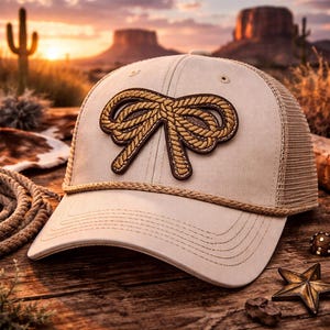 May include: Tan trucker hat with a brown rope bow patch. The hat has a mesh back and a curved brim. The hat is sitting on a wooden surface with a desert landscape in the background. A star-shaped metal object is visible.