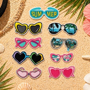 May include: A collection of embroidered sunglasses patches in various shapes and colors, including heart and cat-eye styles, with the word "SUMMER" on one. The patches are arranged on a sandy surface with seashells, a starfish, and tropical flowers.