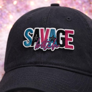 May include: Black baseball cap with the word "SAVAGE" in white, black, teal, and pink lettering. The word "bitch" is written in pink script below the word "SAVAGE". The cap has a curved brim and a fabric strap closure.