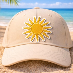May include: A beige baseball cap with a yellow sun patch. The sun design has a yellow center and yellow rays with white outlines. The cap is on a sandy beach with the ocean in the background.