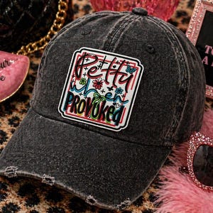 May include: A distressed black baseball cap with a colorful patch that reads "Pretty When Provoked." The patch features a mix of fonts and floral accents. The cap has a curved brim and a worn appearance.