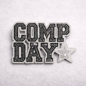 May include: A decorative pin with the words "COMP DAY" in black, outlined with silver rhinestones. A silver sequined star is next to the text. The pin is on a white background.