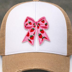 May include: A trucker hat with a white front panel and a brown mesh back. The front features a pink bow patch decorated with red strawberries. The brim is distressed.