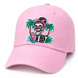 May include: Pink baseball cap featuring an embroidered patch of a skeleton wearing heart-shaped sunglasses and a pink bow, sipping a tropical drink. The design includes palm trees and a black bun. A casual accessory.