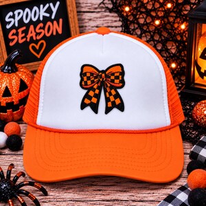 May include: A white and orange trucker hat featuring a central embroidered patch of a bow with a black and orange checkered pattern. The hat has a solid white front panel, an orange brim, and orange mesh side panels with a matching orange cord.