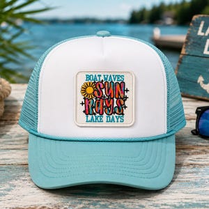 May include: A light blue and white trucker hat featuring a square patch on the front panel. The patch displays a yellow sun icon and the colorful text "BOAT WAVES SUN RAYS LAKE DAYS" in a mix of blue, pink, and orange hues with black outlines.