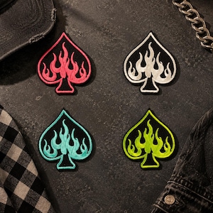 May include: Four embroidered spade-shaped patches featuring flame designs. Each patch has a black background with a distinct flame color: one pink, one white, one turquoise, and one lime green. The patches are arranged in a two-by-two grid pattern.