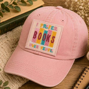 May include: A dusty rose baseball cap with a square patch that reads "I Prefer Books Over People" in colorful embroidered letters. The cap is made of a soft fabric and has a curved brim. A perfect accessory for book lovers.