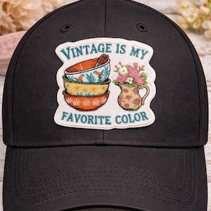 May include: Black baseball cap with a patch that reads "Vintage is my favorite color." The patch features a stack of decorative bowls and a floral pitcher. The bowls have floral patterns in orange, yellow, and blue.