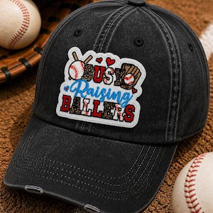 May include: Black baseball cap with a distressed finish. The cap features a patch with the text "Busy Raising Ballers" in red, blue, and white. Baseball and glove graphics are also on the patch. Two baseballs and a glove are in the background.