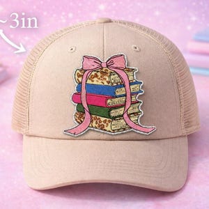 May include: A beige trucker hat with a stack of books embroidered on the front. The books are in various colors, and a pink bow is tied around them. The hat is approximately 3 inches in height.