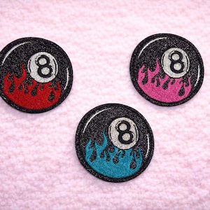 May include: Three round patches featuring a black glitter background with a white number 8. Each patch has flames in red, pink, and blue. The patches are likely for clothing or accessories.