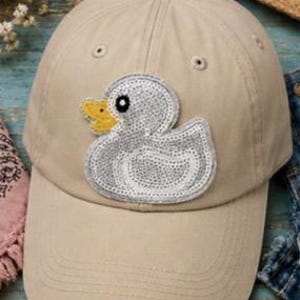 May include: Tan baseball cap featuring a sequined duck applique with a yellow beak. The cap has a curved brim and adjustable strap. The hat is a neutral color and suitable for casual wear.