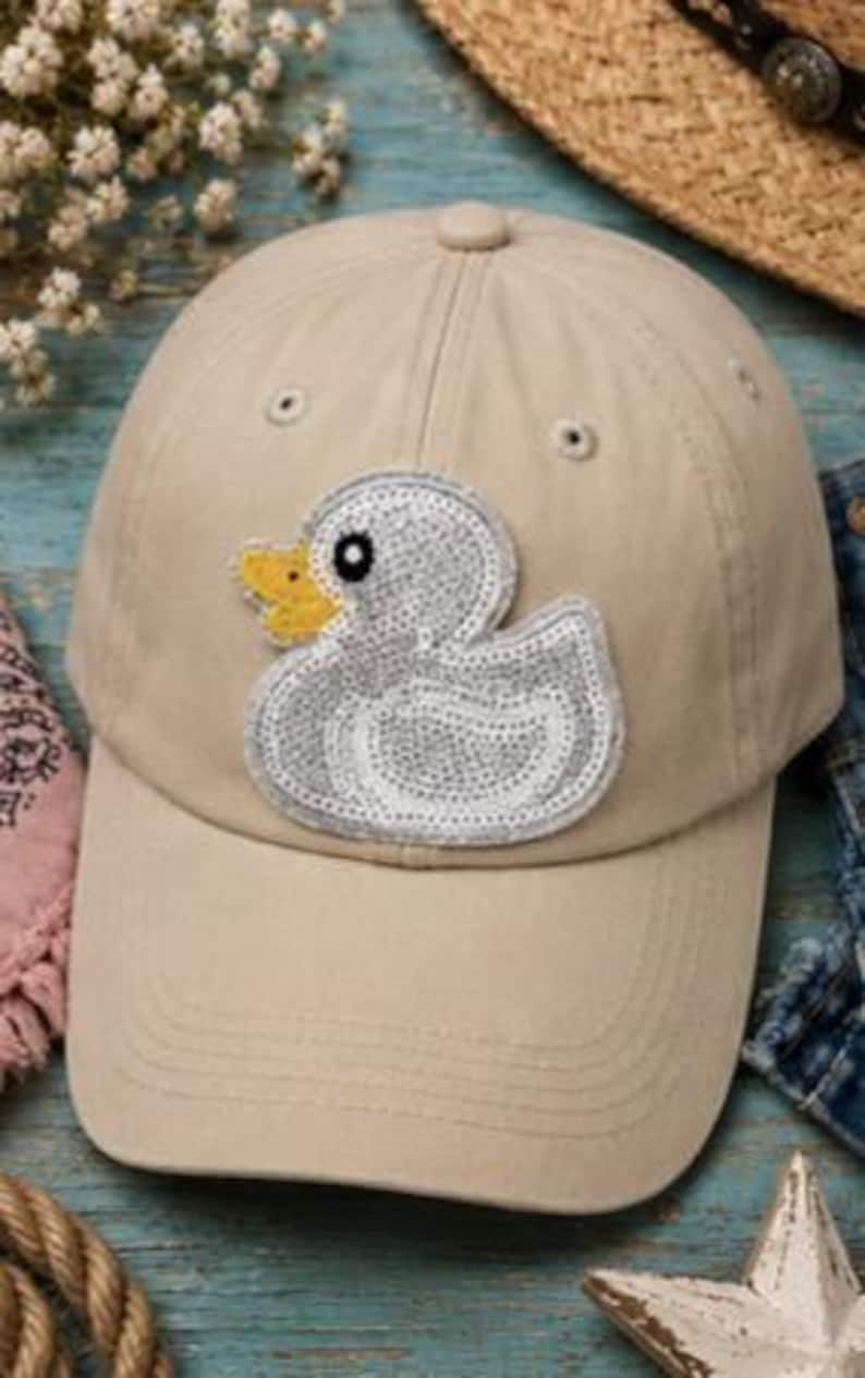 May include: Tan baseball cap featuring a sequined duck applique with a yellow beak. The cap has a curved brim and adjustable strap. The hat is a neutral color and suitable for casual wear.