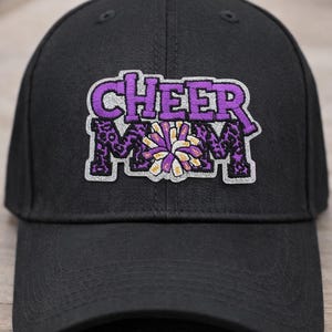 May include: Black baseball cap with the embroidered text "CHEER MOM" in purple and white. The letters have a leopard print design. A purple and white pom-pom is in the center of the text. The hat is a classic baseball cap style.