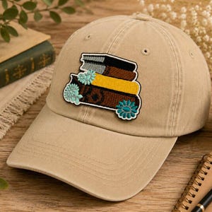 May include: A tan baseball cap with a patch featuring a stack of books in brown, yellow, and black, accented with teal floral details. The cap is made of a soft fabric and has a curved brim.