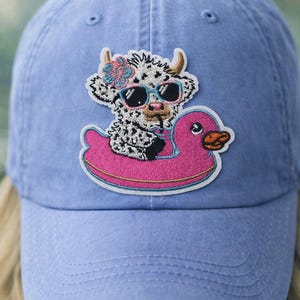 May include: A light blue baseball cap featuring an embroidered patch of a cow wearing sunglasses and a flower, floating on a pink duck-shaped inflatable. The cap has a curved brim and a fabric strap.