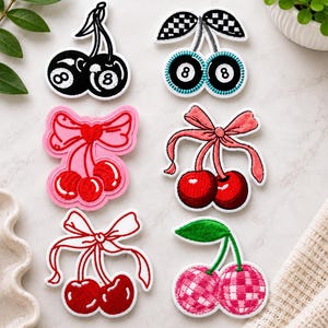 May include: Six embroidered patches featuring cherry motifs. Two patches depict cherries as black 8-balls, one with a checkered leaf. Two patches show red cherries with pink or red bows. One patch has red cherries with a white bow, and one has pink checkered cherries.
