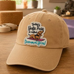 May include: A tan baseball cap with a curved brim. The cap features a patch with the text "Today's Yoga Pose is called Downward Spiral" and a cartoon dachshund doing yoga. The cap is made of a soft, durable material.