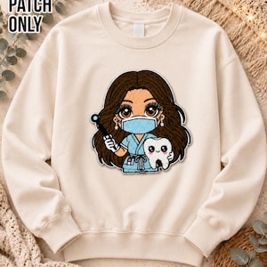 May include: A beige crewneck sweatshirt featuring a central embroidered patch of a cartoon dental professional with long brown hair, wearing a blue scrub top and a light blue face mask, holding a dental tool and a smiling white tooth character.