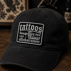 May include: Black baseball cap with a rectangular patch that reads "tattoos because (life) is hard and I deserve stickers". The cap is made of a soft, worn material.