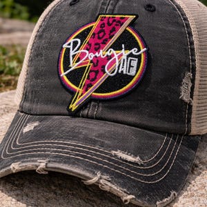 May include: Distressed black baseball cap with a tan mesh back. The cap features a circular patch with the word "Bougie" in white script, a pink leopard print lightning bolt, and a yellow and purple border.