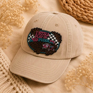 May include: Tan baseball cap with a patch that reads "I Still Play in Mud Puddles." The patch features a checkered flag design and an image of an off-road vehicle. The cap is made of a soft material.