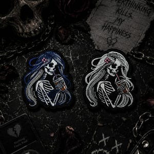 Pu&ograve; includere: Due toppe ricamate raffiguranti scheletri con capelli lunghi e bevande. Uno ha i capelli blu, l'altro bianchi. L'immagine include accessori gotici: un teschio, catene e un biglietto con la scritta "Overthinking Kills My Happiness". Il tema &egrave; dark e gotico.