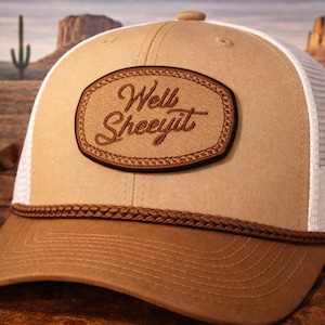 May include: Tan and brown trucker hat with a white mesh back. The hat features a brown leather patch with the words "Well Sheeeyit" in a script font. The brim is brown, and the hat has a braided brown rope detail.
