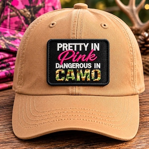 May include: A tan baseball cap with a black patch that reads "PRETTY IN Pink DANGEROUS IN CAMO". The text is in white and pink, with the word "CAMO" in a camouflage pattern. The hat has a curved brim.