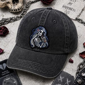 May include: A black, distressed baseball cap with a patch featuring a skeleton with long blue hair drinking from a cup. The cap has a curved brim and a classic six-panel design. The text "Overthinking Kills My Happiness" is visible.