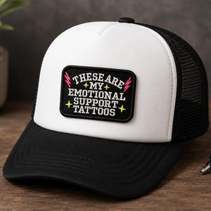 May include: A black and white trucker hat featuring a rectangular patch on the front. The patch is black with white text that reads, "THESE ARE MY EMOTIONAL SUPPORT TATTOOS," accented by small yellow stars and pink lightning bolt graphics.
