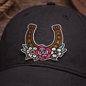 May include: A black baseball cap featuring a floral horseshoe patch. The patch includes a brown horseshoe with a pink rose, white and yellow flowers, and green leaves. The cap is made of a dark fabric.