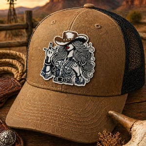 May include: A brown trucker hat with a black mesh back and a detailed patch featuring a cowgirl illustration. The patch shows a cowgirl wearing a hat and making a hand gesture. The hat has a curved brim and a button on top. The background shows a sunset and a windmill.