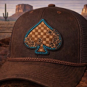 May include: Brown trucker hat with a spade-shaped patch. The patch features a checkered pattern, floral accents, and turquoise-colored beading. The hat has a mesh back and a rope detail along the brim. A turquoise-colored accessory is attached to the side.