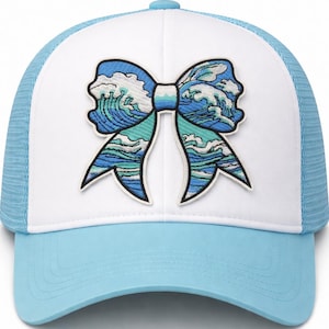 May include: A trucker hat with a white front panel and a light blue brim and mesh back. The hat features an embroidered bow design with ocean wave patterns in shades of blue and teal.